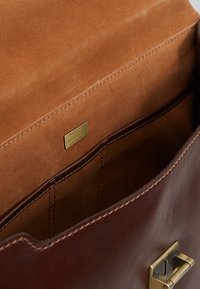 Interior de bolsa de cuero marrón con forro de ante beige, cuenta con una placa en tono dorado y compartimentos para la organización. Acentuada con herrajes dorados.
