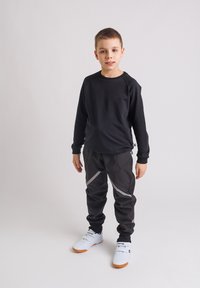 Chłopiec stoi w czarnej koszulce z długim rękawem, czarnych spodniach z odblaskowymi paskami i białych trampkach na jednolitym jasnym tle.
