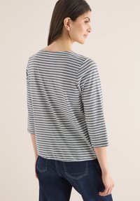 Femme de profil portant un haut gris et blanc à manches trois-quarts rayé et un jean bleu foncé sur un fond uni.