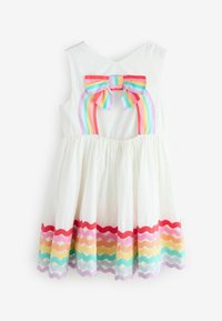 Vestido blanco sin mangas para niña con lazo arcoíris en la parte trasera y ribete de ondas arcoíris a juego cerca del dobladillo.