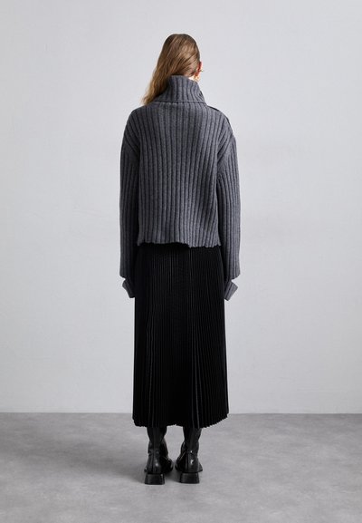 Suéter cinza com gola alta canelada combinado com uma saia midi plissada preta. Botas pretas chunky completam o visual, visíveis por trás.