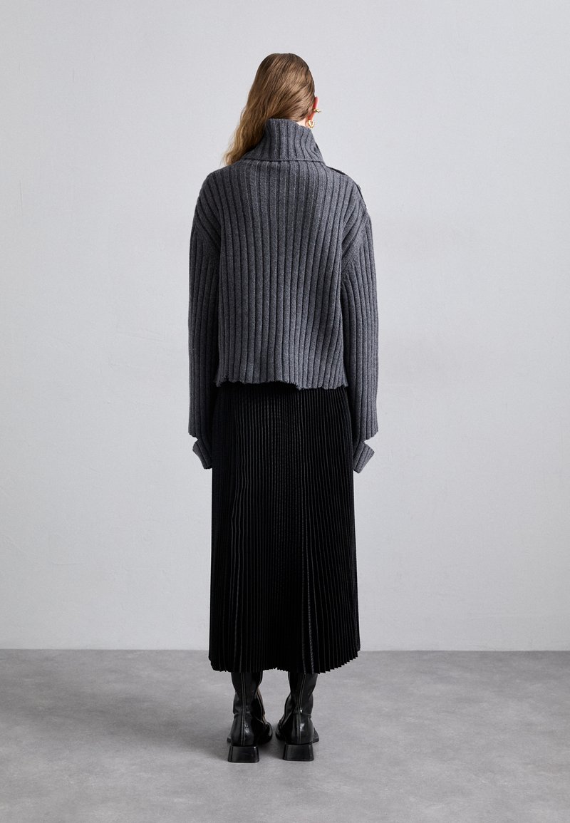 Grijze geribde coltrui gecombineerd met een zwarte plissé midi-rok. Chunky zwarte laarzen maken de outfit compleet, zichtbaar vanaf de achterkant.