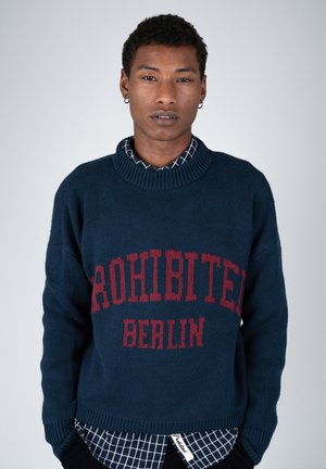 Jeune personne aux cheveux courts et bouclés portant un pull bleu marine avec un texte rouge "PROHIBITED BERLIN" par-dessus une chemise à carreaux.