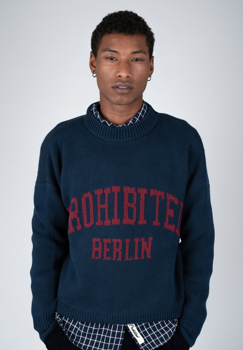 Jeune personne aux cheveux courts et bouclés portant un pull bleu marine avec un texte rouge "PROHIBITED BERLIN" par-dessus une chemise à carreaux.
