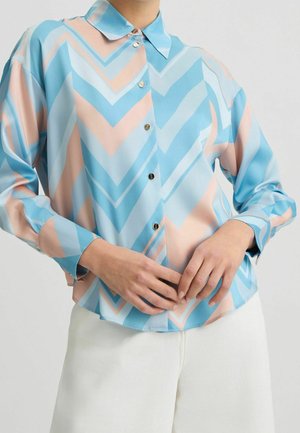 Koszula zapinana na guziki z pasami w zygzak w pastelowych odcieniach niebieskiego, różowego i białego, noszona z białymi spodniami, ręce poprawiające dół koszuli.