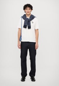 Balti kokvilnas T-krekls ar mazu izšūtu dizainu, tumši zilā rakstaina džemperis sasieta ap kaklu, tumši džinsi un melni eleganti apavi.