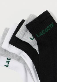 Pilha de seis meias até ao tornozelo nas cores preta, cinza e branca com logos verdes da Lacoste perto da bainha, dispostas sobre um fundo branco.