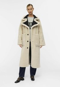 Übergroßer beiger Schaffellmantel mit schwarzem Kragen, vorderen Knöpfen und zwei großen Taschen; getragen über einem grauen Hemd und blauen Jeans.