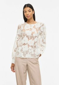 Femme portant un chemisier en dentelle blanche transparente à motifs floraux avec de longues manches bouffantes et un pantalon beige, debout devant un fond uni.