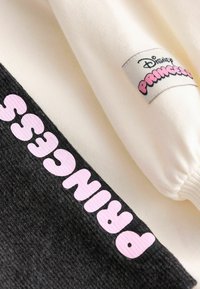 Balts un melns kapucis, uz kura rakstīts "PRINCESE" rozā krāsā, ar nelielu Disney etiķeti augšējā šuvē. Mīksts kokvilnas maisījuma materiāls.