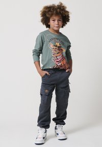 Zelena majica s dugim rukavima s pisanom slikom dinozaura, u kombinaciji s tamnim cargo hlačama. Footwear sadrži bijele i mornarsko plave detalje.