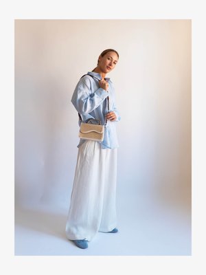 Světle modrá košile s dlouhým rukávem na knoflíky, bílé kalhoty s širokými nohavicemi, modré slip-on boty a malá béžová a hnědá kabelka s texturovaným designem.