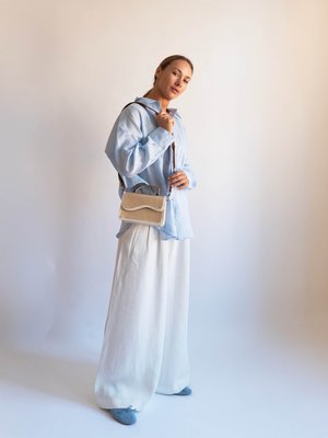 Woman in light blue shirt, white wide-leg pants, blue flats, holding beige crossbody bag with woven texture against plain backdrop.