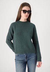Pull vert texturé avec un col rond et un ourlet côtelé. Porté avec un jean bleu et des lunettes de soleil noires.