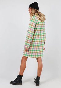 Robe chemise à carreaux verte, violette, marron et jaune, oversized, avec des manches longues, des poignets à boutons et un ourlet arrondi, portée avec des bottes noires.
