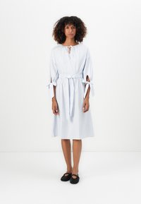 Vestido de algodão riscado com decote redondo, detalhes de laço na cintura e nas mangas, e um corte solto, combinado com sapatos pretos com tachas.