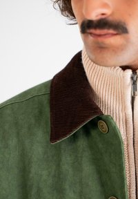 Grøn suedjacket med en brun corduroy-krave. Har en lynlåset beige ribbestrikket sweater under. Nærbillede, der fremhæver teksturer.