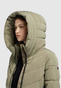 Chaqueta tipo puffer en verde oliva con una gran capucha, diseño acolchado y cremallera negra, con un parche de logo en el lateral y una textura ligera.