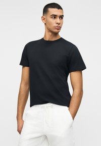 Νέος άνδρας που φορεί απλό μαύρο κοντομάνικο μπλουζάκι και λευκό παντελόνι με κορδονάκι, κοιτάζοντας προς τα αριστερά με τα χέρια στις τσέπες.