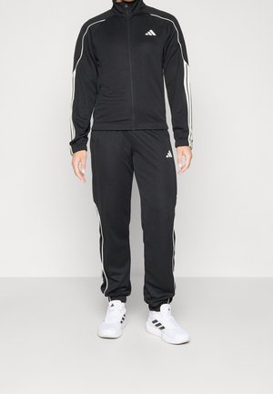 Osoba ubrana w czarny dres Adidas z białymi paskami i logo, w połączeniu z białymi sneakersami Adidas, stojąca na jednolitym tle.