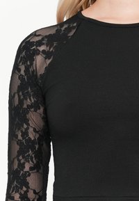 Haut noir ajusté, avec de longues manches en dentelle transparente à motif floral et un col rond. Texture douce.