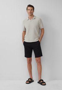 Hellgraues, strukturiertes Poloshirt mit kurzen Ärmeln, kombiniert mit schwarzen Shorts und schwarzen Sandalen, Hände in den Taschen vor einem schlichten Hintergrund.