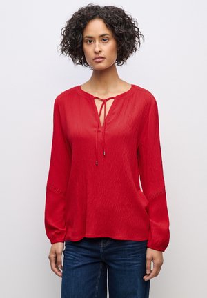 Blouse rouge à manches longues avec un col rond et un détail noué. Tissu texturé et accouts en point ouvert sur les poignets.