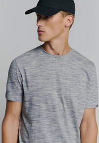 Grijze en blauwe gespikkelde T-shirt met korte mouwen en een ronde hals, gedragen onder een zwarte pet. De stof lijkt textuur te hebben met subtiele variaties.