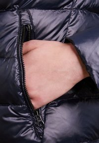 Chaqueta acolchada de color azul marino con un bolsillo con cremallera, que muestra una textura lisa y brillante, con una mano apoyada dentro del bolsillo.