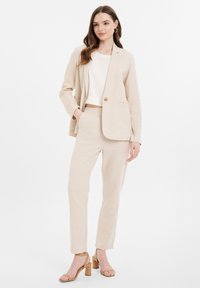 Béžový ľanový blazer s ladiacimi nohavicami, s jednoduchým zapínaním na gombík, vreckami a krátkym bielym topom; model má na sebe béžové sandále na blokovom podpätku.