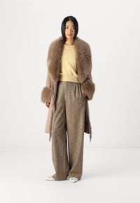 Καφέ παλτό με γούνα στον γιακά και τα μανίκια, κίτρινο πουλόβερ και καρό παντελόνι φαρδιών ποδιών. Γυαλιστερά λευκά παπούτσια με χρυσές λεπτομέρειες.