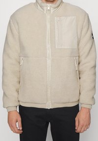 Krämfärgad fleecejacka med dragkedja fram, ribbade muddarna och nederkant. Har ett texturerat tyg och en bröstficka i ett slätt material.