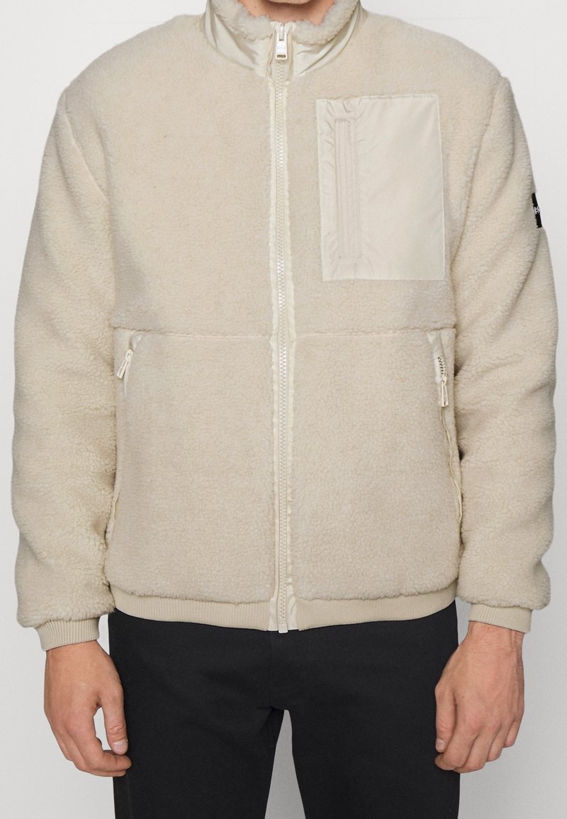 Krämfärgad fleecejacka med dragkedja fram, ribbade muddarna och nederkant. Har ett texturerat tyg och en bröstficka i ett slätt material.