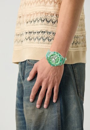 Montre numérique translucide verte avec plusieurs cadrans, bracelet en plastique texturé, présentée sur une main, associée à un top en tricot beige.