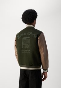 Chaqueta universitaria verde con mangas de cuero marrón, con bordado de "Casa de los Poetas" y un gráfico de un edificio en la parte posterior, cuello acanalado a rayas.
