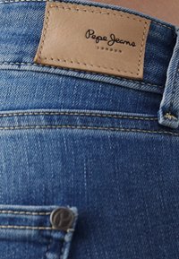 Denimjeans har en ljusblå färg, en läderlapp med "Pepe Jeans London"-logotypen och en metallknapp med en logotyp.