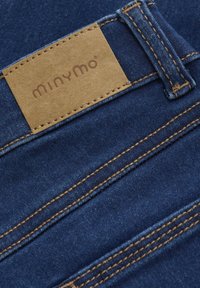 Traperice s kožnom oznakom boje pijeska s natpisom "minymo." Imaju kontrastne šavove i glatku teksturu u tamnoplavoj tkanini.
