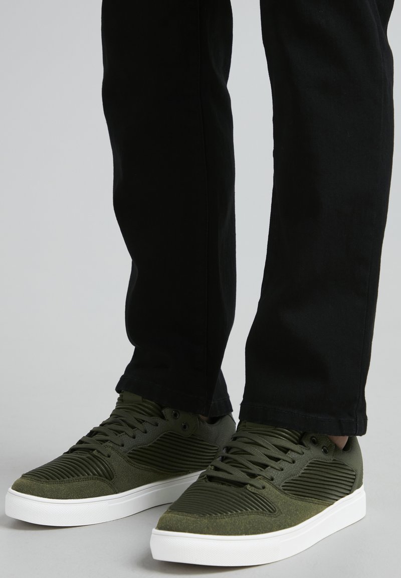 Gröna sneakers med texturerat meshtyg, ribbat designaccent, rund tå och vit gummisula, matchat med svarta byxor.