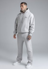 Lichtgrijze hoodie met rits, voorzakken en een logo. Gecombineerd met bijpassende joggingbroek met logo op de heup. Witte sneakers maken de look compleet.