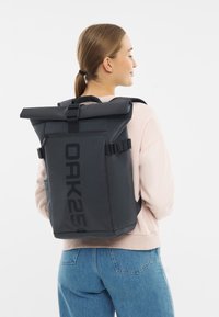 Mochila negra tipo roll-top con correas ajustables, que cuenta con un acabado liso e impermeable y un gran logo impreso en el lateral.