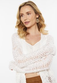 Cardigan bianco, lavorato a maglia, con un design in pizzo, maniche lunghe e apertura frontale. Indossato sopra un top leggero, caratterizzato da dettagli texture.