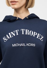 Nærbilde av en person som har på seg en marineblå hettegenser med hvit brodert tekst "SAINT TROPEZ" og "MICHAEL KORS" på forsiden.