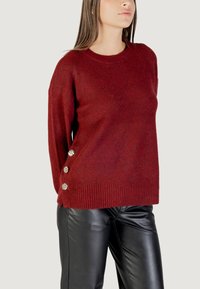 Maglione in maglia borgogna con orlo e polsini a coste, caratterizzato da tre bottoni decorativi in tonalità argentata sul lato sinistro. Indossato con pantaloni in pelle nera.