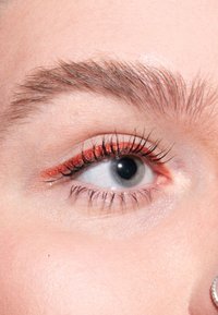 Nærbilde av et øye med myk hud, mørk pupill og lysfarget iris. Lys rød eyeliner fremhever vippene og forbedrer øyets form.