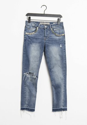 Jeans di denim blu con ginocchio strappato, orli sfrangiati e decorazioni di strass lungo le tasche anteriori, appesi a una gruccia nera.