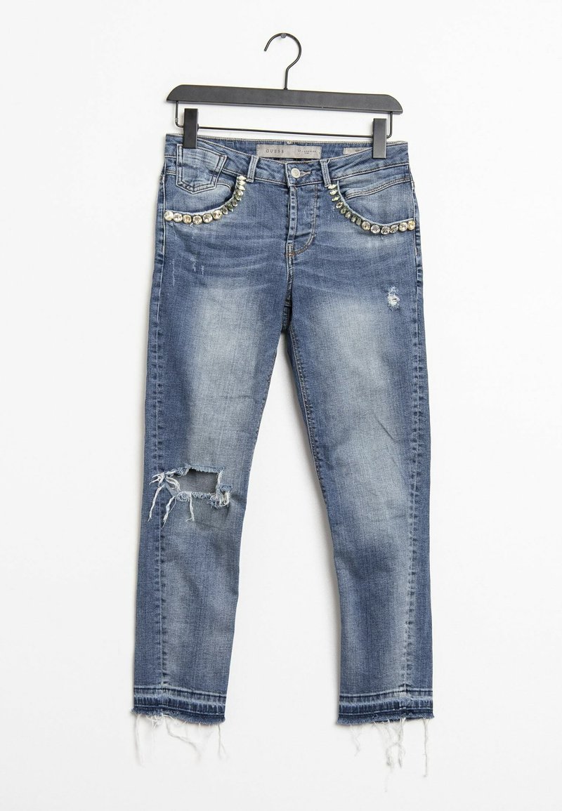 Jean en denim bleu avec genoux déchirés, ourlets effilochés et ornements en strass le long des poches avant, suspendu à un cintre noir.