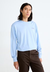 Camiseta de manga larga de color azul claro con cuello redondo, con un pequeño logo morado de "nike" en el pecho, combinada con pantalones marrón oscuro.