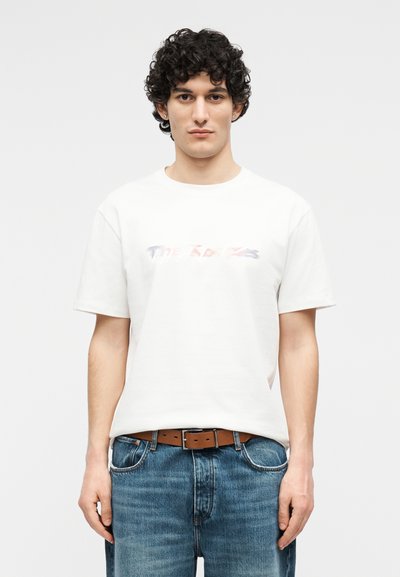 Νέος άνδρας με σγουρά σκούρα μαλλιά, που φοράει λευκό κοντομάνικο μπλουζάκι και μπλε τζιν με καφέ ζώνη, στέκεται μπροστά σε απλό φόντο.