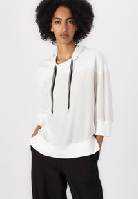 Weißer Kapuzenpullover mit lässiger Passform, ausgestattet mit schwarzen Kordeln und 3/4-Ärmeln, kombiniert mit schwarzen Hosen. Leichter, weicher Stoff.