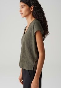 Olive green, V-neck top with short sleeves and textured fabric. Features a relaxed fit and side slits for ease of movement.